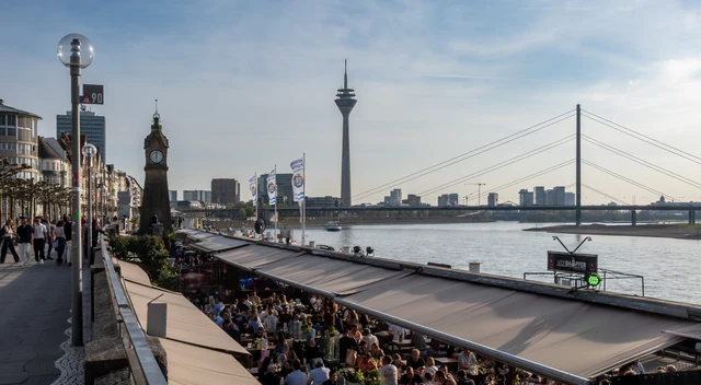 Belebte Gastronomieterrassen an der Rheinuferpromenade in Düsseldorf mit Pegeluhr, Rheinturm und Rheinkniebrücke