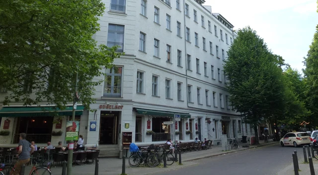 Helles Berliner Eckhaus mit Straßencafé und grünen Bäumen am Kollwitzplatz in Prenzlauer Berg